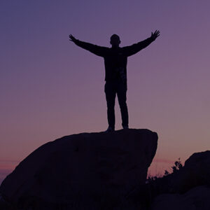 Silhouette of person with out stretched arms standing on a rock at dusk