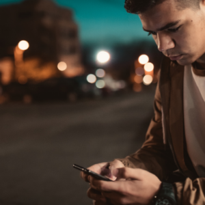 A teenager looking at their smart phone on a street at night time.
